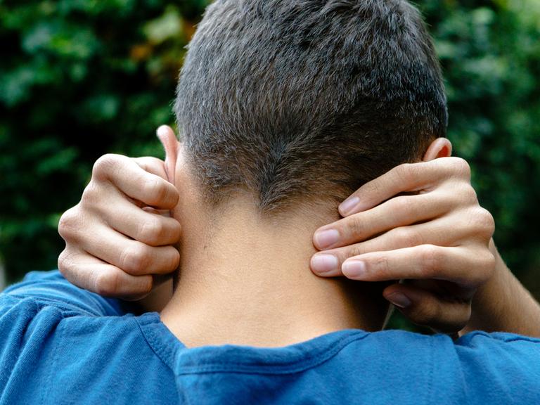 Ein autistischer junger Mann, fotografiert von hinten, er hält sich die Ohren zu.