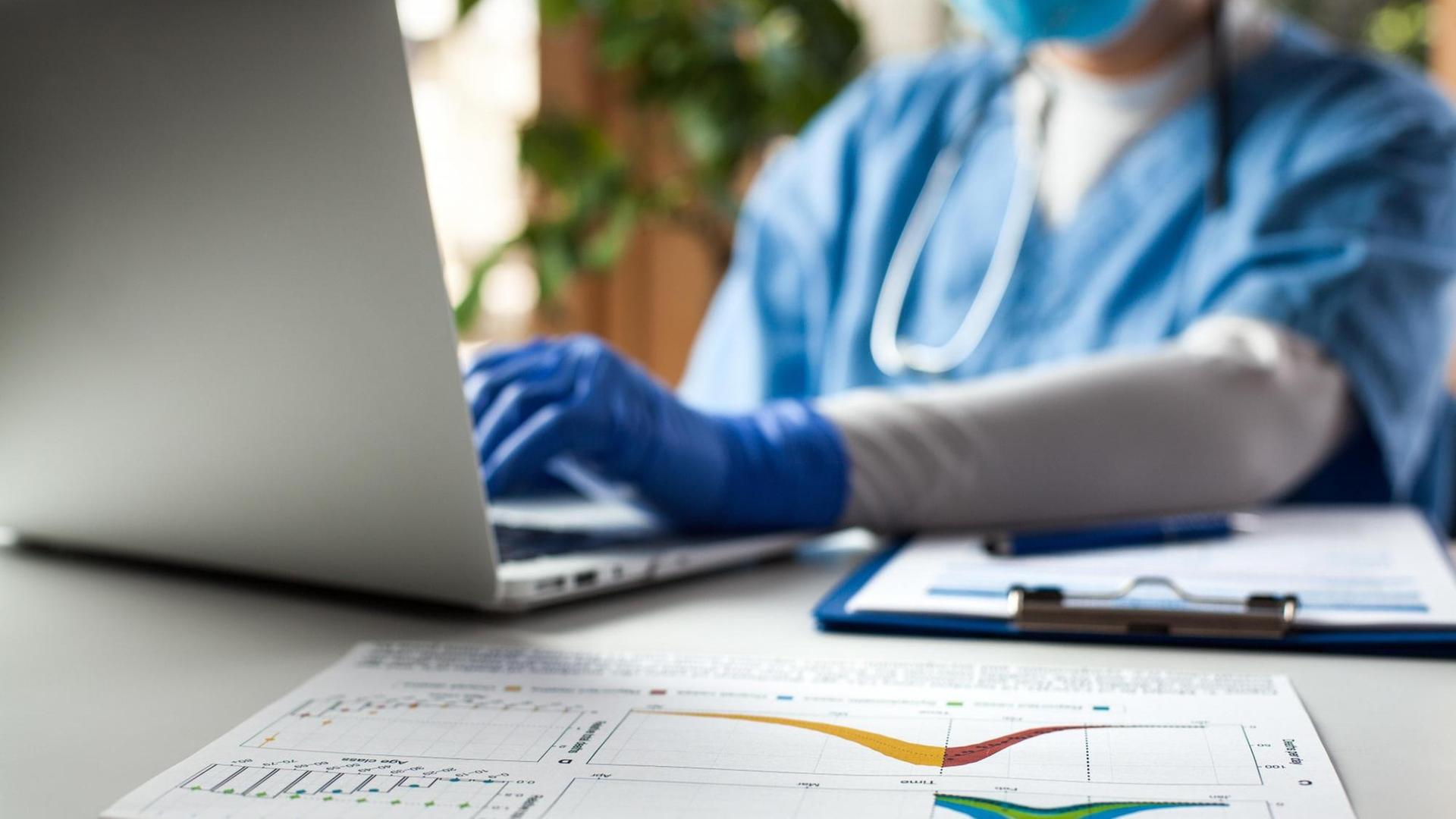 Ein Mediziner arbeitet an einem Laptop, daneben liegen Ausdrucke von Statistiken.