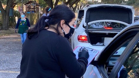 Eine junge Frau mit schwarzem Pulli und Mundschutz steht an dem heruntergelassenen Fenster eines Autos und macht sich Notizen.