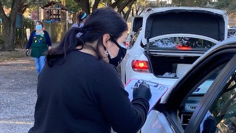 Eine junge Frau mit schwarzem Pulli und Mundschutz steht an dem heruntergelassenen Fenster eines Autos und macht sich Notizen. Eine junge Frau mit schwarzem Pulli und Mundschutz steht an dem heruntergelassenen Fenster eines Autos und macht sich Notizen.