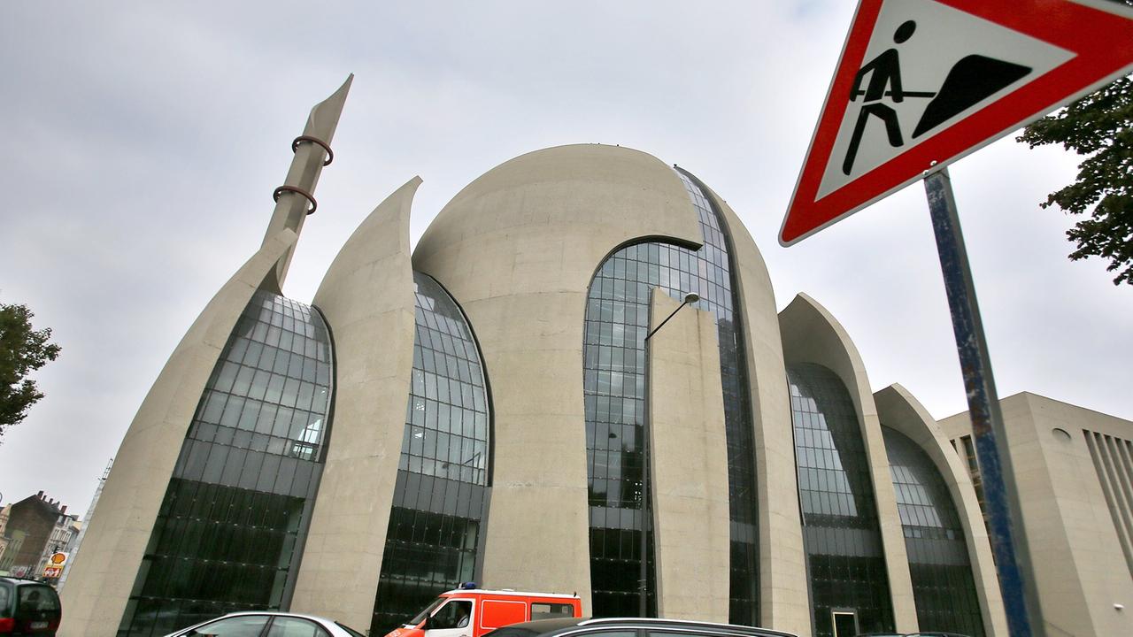 Ansicht der Kölner Zentralmoschee, eines geschwungenen Gebäudes mit Kuppel aus Beton und Glas, davor Autos auf einer Straße und ein Baustellenschild Ansicht der Kölner Zentralmoschee, eines geschwungenen Gebäudes mit Kuppel aus Beton und Glas, davor Autos auf einer Straße und ein Baustellenschild