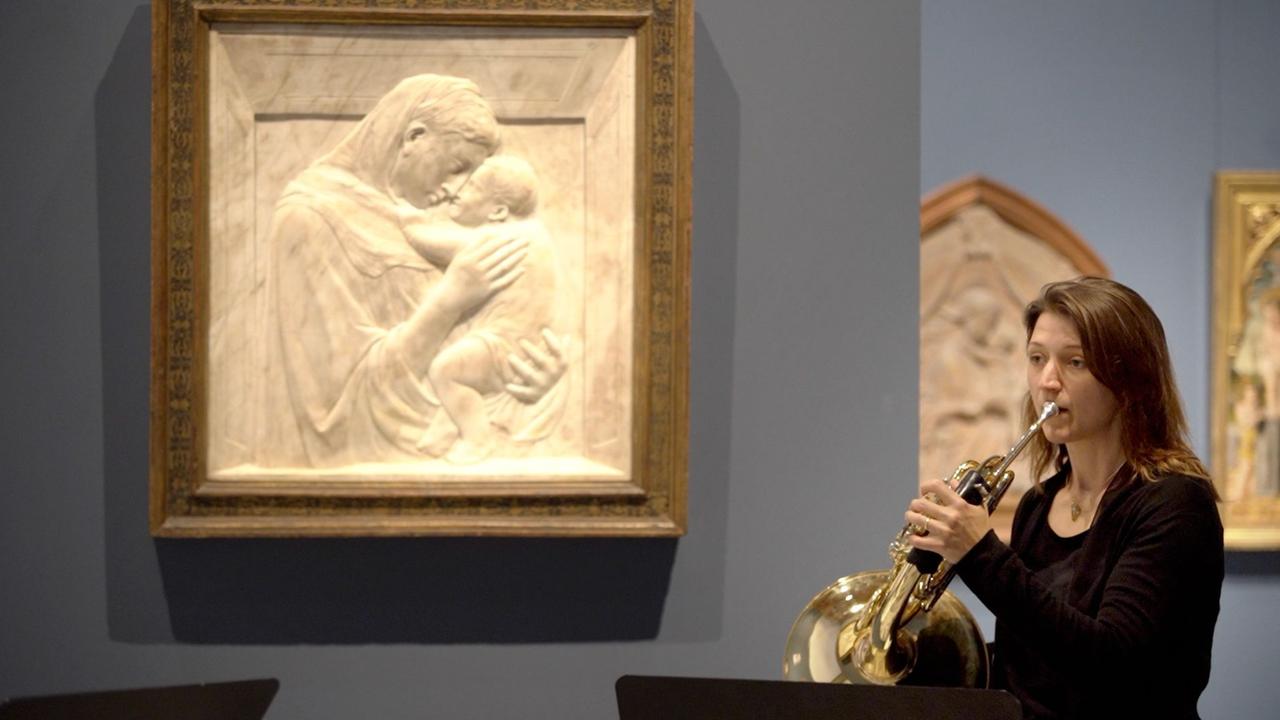Eine Hornistin spielt inmitten eines Museums, neben ihr ein Relief, das eine Frau vertieft in die Umarmung eines Kindes zeigt. Eine Hornistin spielt inmitten eines Museums, neben ihr ein Relief, das eine Frau vertieft in die Umarmung eines Kindes zeigt.