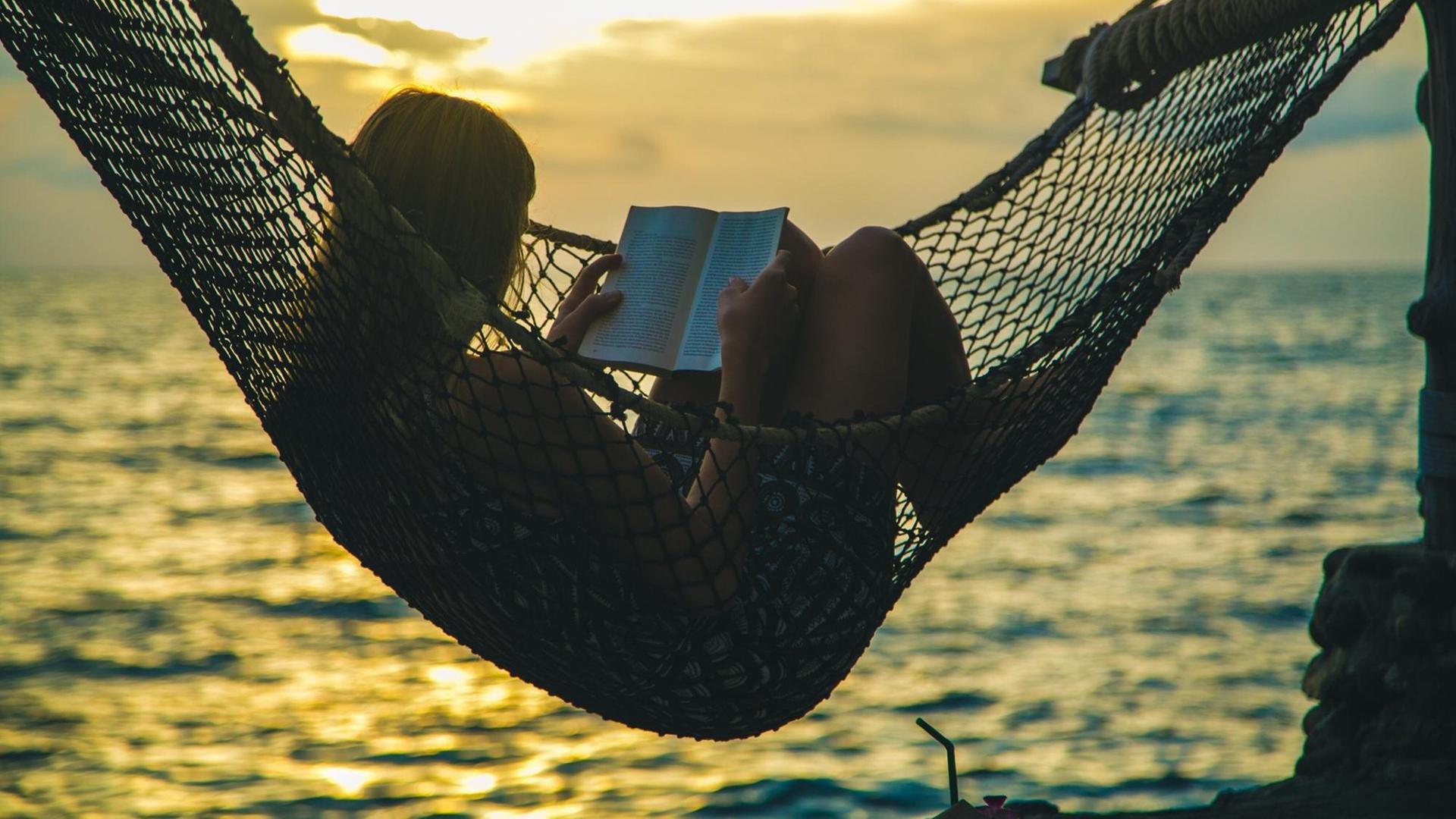 Eine Frau liegt in der Hängematte und liest ein Buch. Im Hintergrund das Meer bei Abendstimmung.