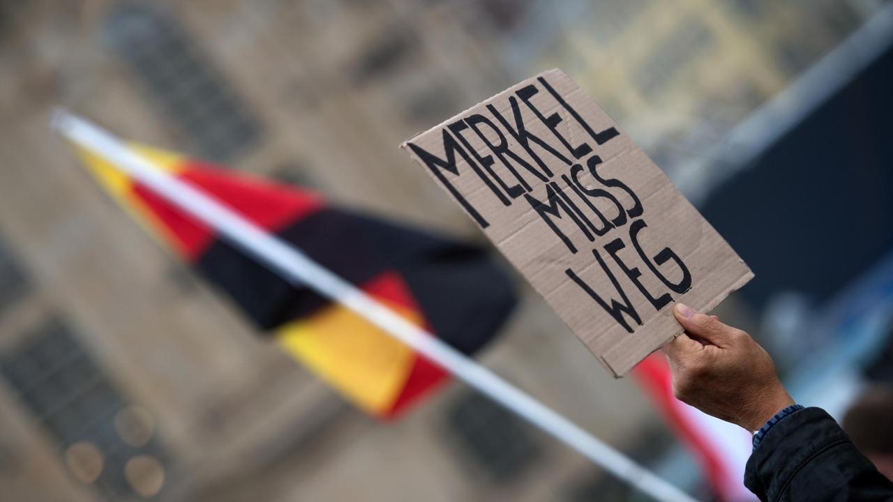 Eine Hand hält ein Schild mit der Aufschrift "Merkel muss weg" in die Höhe, dahinter eine deutsche Flagge. Eine Hand hält ein Schild mit der Aufschrift "Merkel muss weg" in die Höhe, dahinter eine deutsche Flagge.