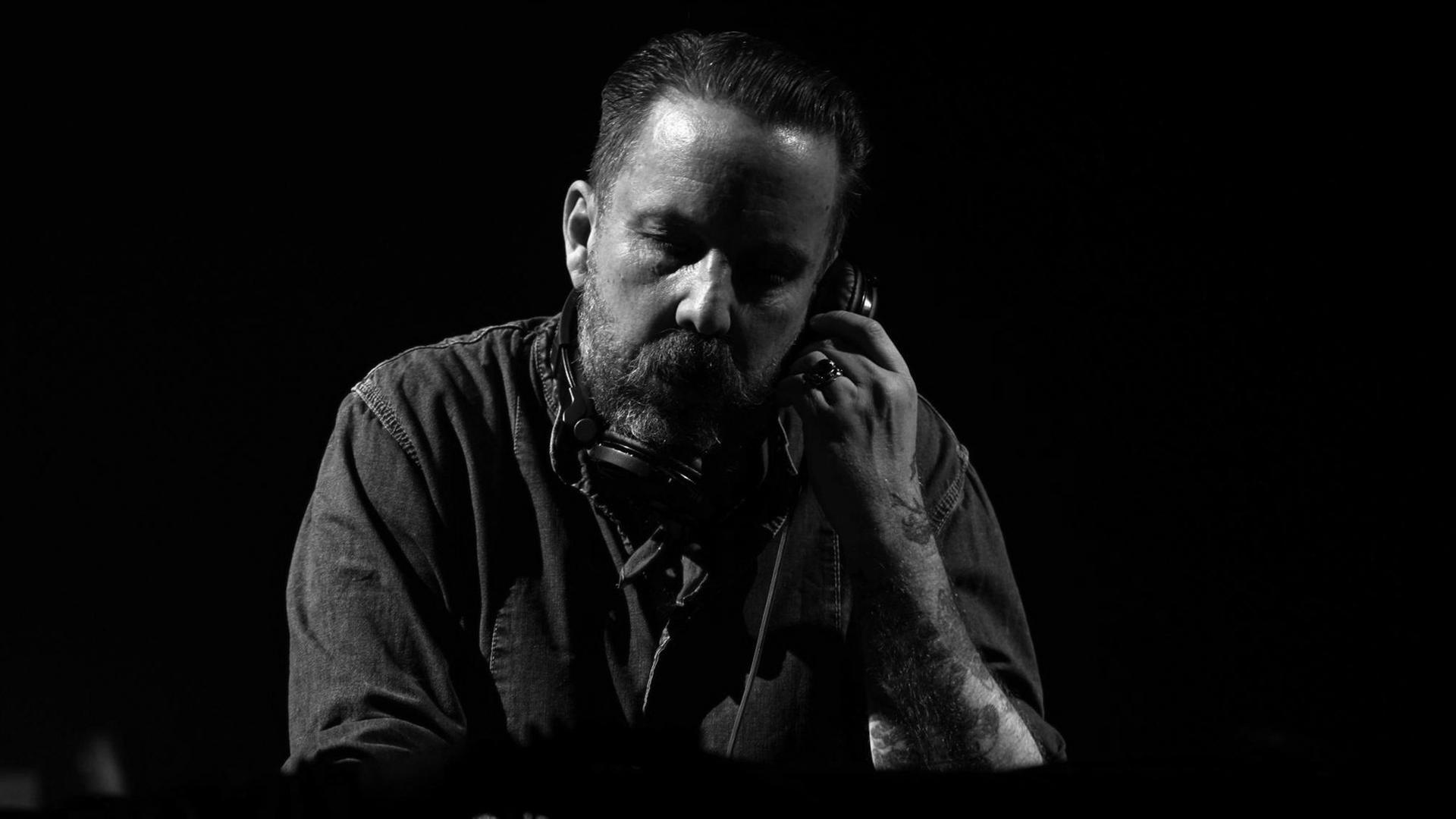 DJ Andrew Weatherall vor dunklem Hintergrund am Mischpult mit Kopfhörern um den Hals. Er hält sich den einenKopfhörer an sein linkes Ohr. DJ Andrew Weatherall vor dunklem Hintergrund am Mischpult mit Kopfhörern um den Hals. Er hält sich den einenKopfhörer an sein linkes Ohr.