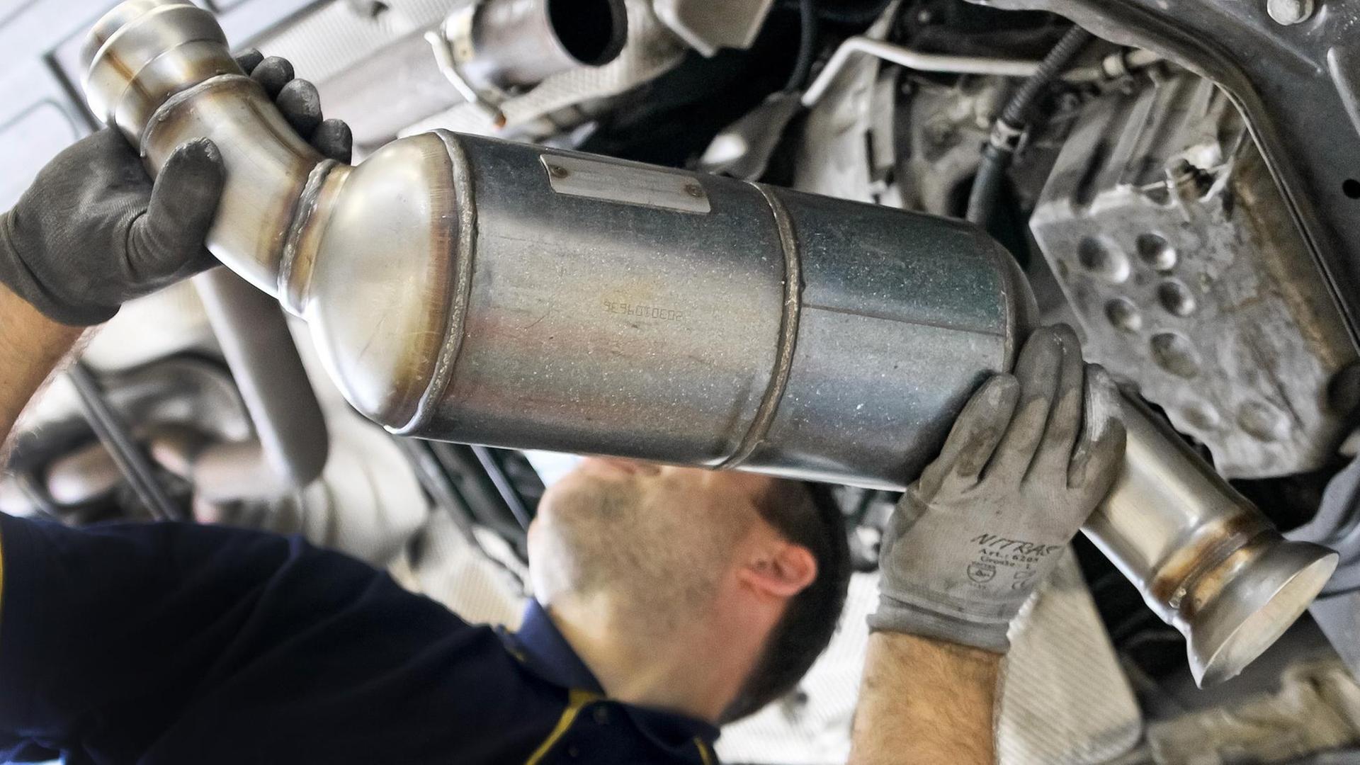 Man sieht, wie der Mechaniker den silber-metallenen Filter von unten in die Unterseite des Fahrzeugs hebt. Er trägt Arbeitshandschuhe. Der Filter verdeckt sein Gesicht. Man sieht, wie der Mechaniker den silber-metallenen Filter von unten in die Unterseite des Fahrzeugs hebt. Er trägt Arbeitshandschuhe. Der Filter verdeckt sein Gesicht.