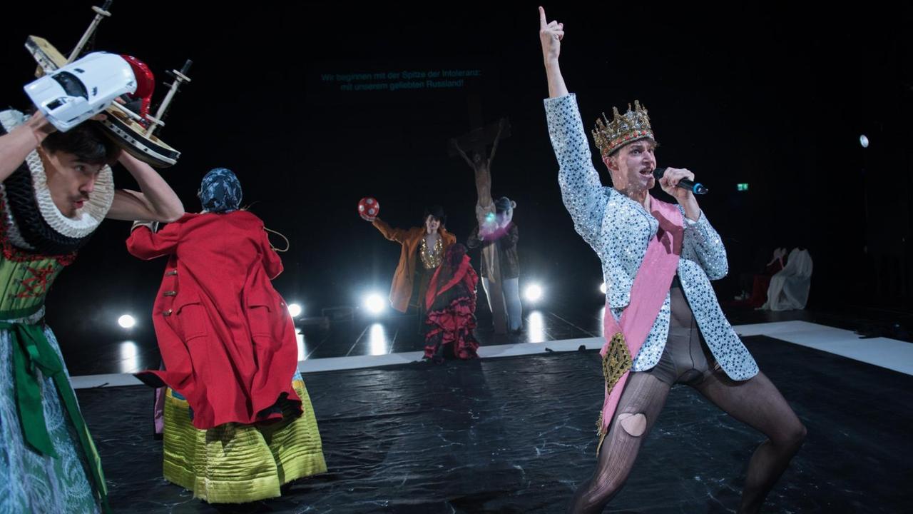 Schauspieler des Schauspiel Stuttgart auf der Bühne in bunten Kostümen. Ein Schauspieler am Bühnenanfang trägt eine Krone und reckt seinen Arm nach oben. Er hält ein Mikrofon und spricht. Schauspieler des Schauspiel Stuttgart auf der Bühne in bunten Kostümen. Ein Schauspieler am Bühnenanfang trägt eine Krone und reckt seinen Arm nach oben. Er hält ein Mikrofon und spricht.
