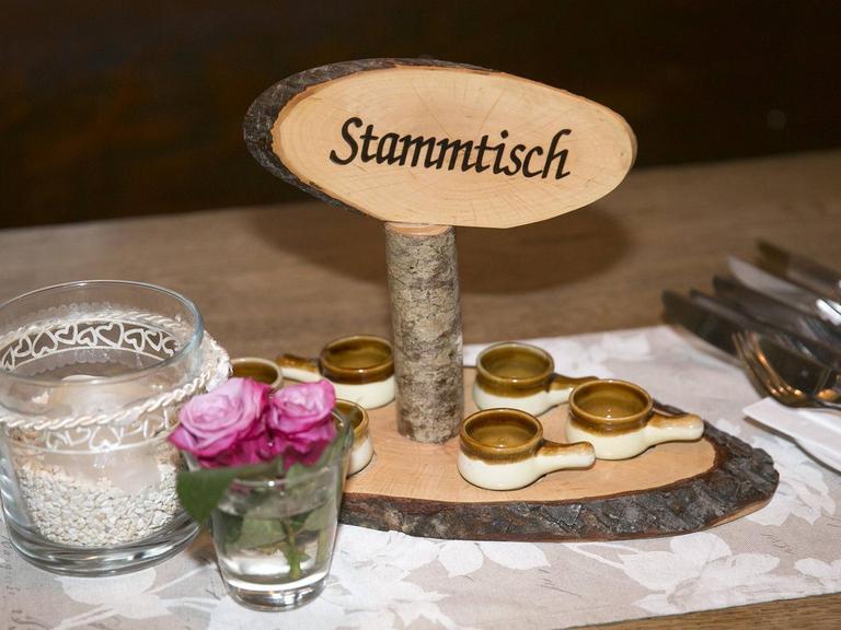 Zu sehen ist die Tischdeko in einem Lokal. Auf dem Tisch stehen Besteck, eine kleine Blumenvase und ein Holzschild in dem "Stammtisch" eingraviert wurde.