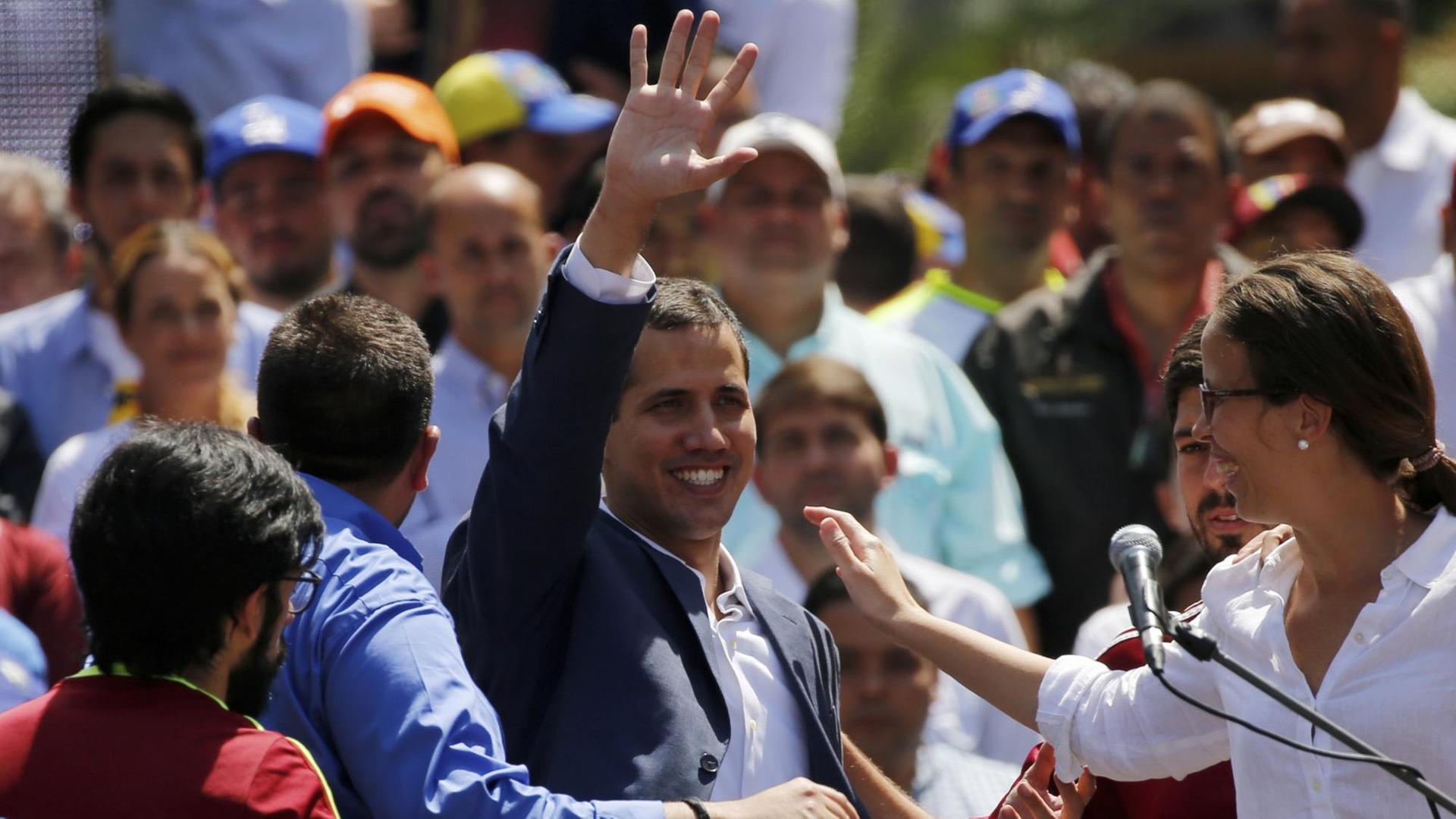 Guaidó steht lächelnd in einer Menschenmenge und winkt; neben ihm steht ein Mikrofon. Guaidó steht lächelnd in einer Menschenmenge und winkt; neben ihm steht ein Mikrofon.