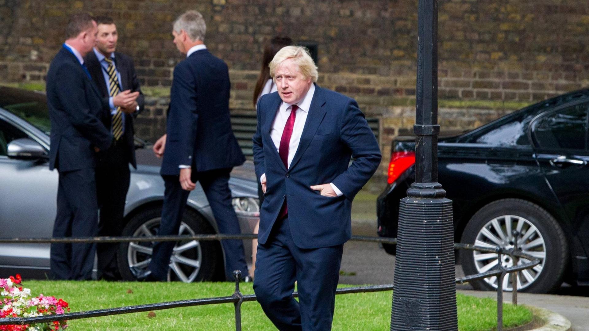 Boris Johnson geht eine Straße entlange, im Hintergrund mehrere Männer und Autos Boris Johnson geht eine Straße entlange, im Hintergrund mehrere Männer und Autos