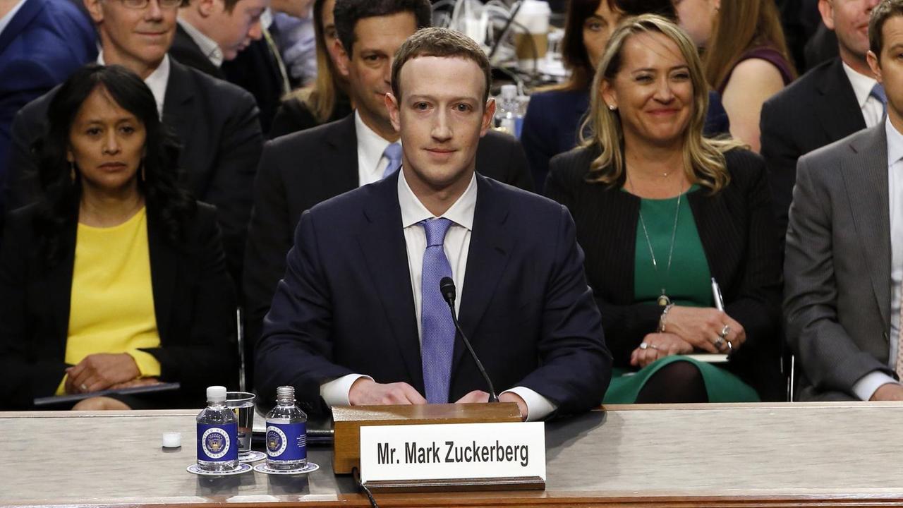 Mark Zuckerberg sitzt an einem Tisch in einem Raum des US-Kongresses, hinter ihm eine Gruppe von Menschen