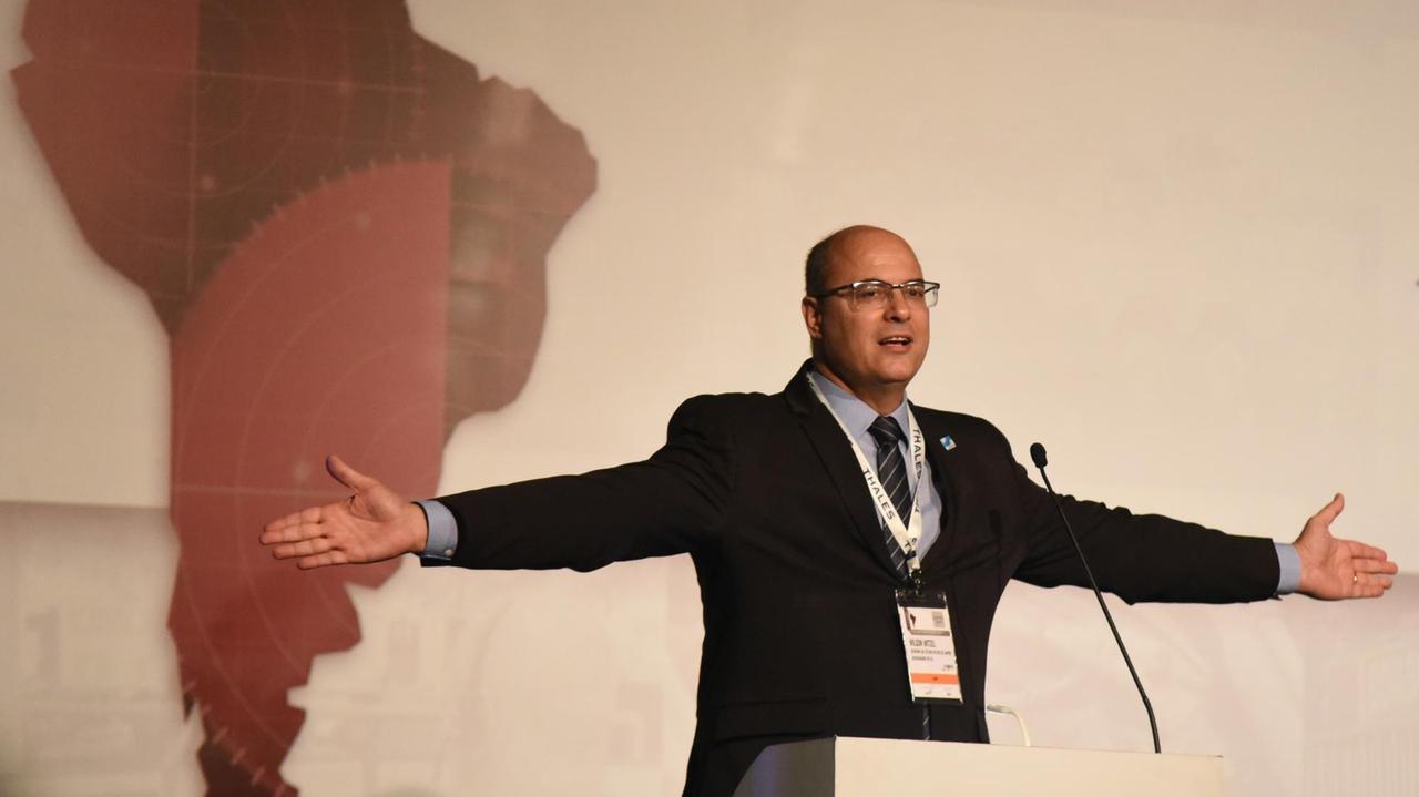 Wilson Witzel, der Gouverneur von Rio de Janeiro, steht hinter einem Podium der LAAD Weapons Fair und spricht mit ausladender Geste. Hinter sich eine stilisierte Karte Brasiliens.