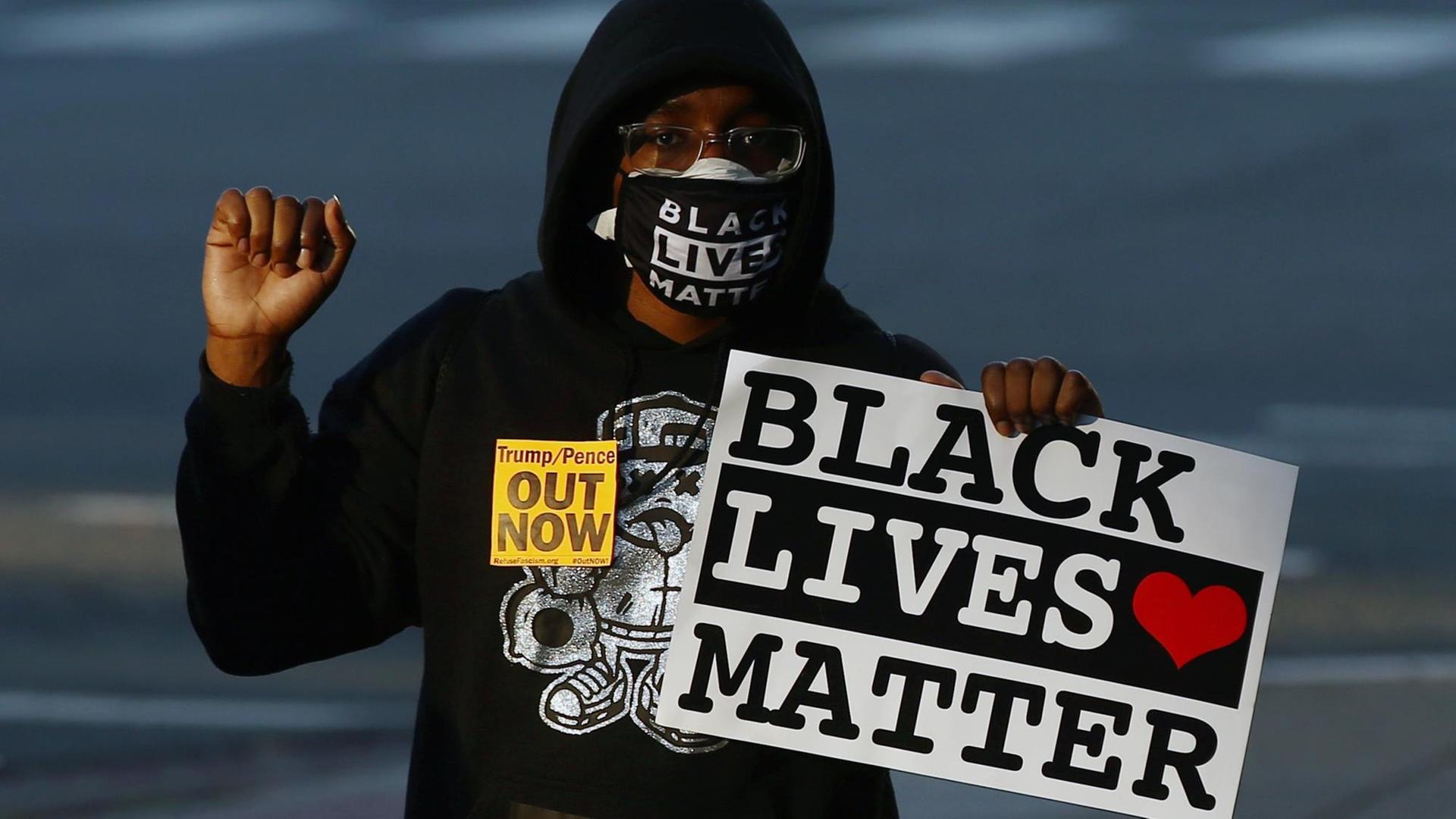 Ein Aforamerikaner mit einem "Black Lives Matter"-Schild in der linken Hand. Der rechte Arm ist nach oben angewinkelt und die Hand zur Faust geballt. Der Mann trägt auch eine Maske mit dem selben Slogan und einen schwarzen Kapuzenpullover.