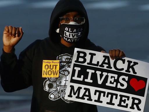 Ein Aforamerikaner mit einem "Black Lives Matter"-Schild in der linken Hand. Der rechte Arm ist nach oben angewinkelt und die Hand zur Faust geballt. Der Mann trägt auch eine Maske mit dem selben Slogan und einen schwarzen Kapuzenpullover.