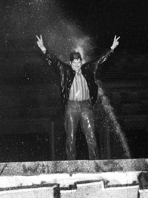 Ein durchnässter Mann steht auf der Berliner Mauer, reckt die Arme in die Höhe und wird dabei vom Strahl eines Wasserwerfers getroffen.