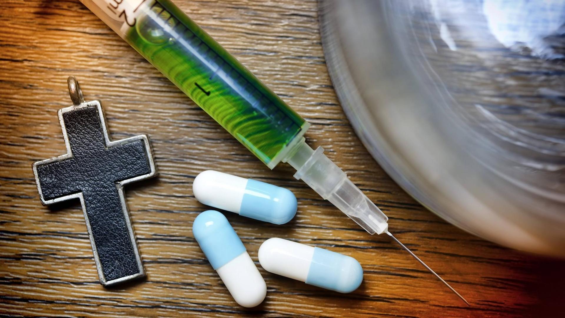 Wasserglas, Kreuz, Tabletten und Spritze liegen auf einem Tisch. Wasserglas, Kreuz, Tabletten und Spritze liegen auf einem Tisch.