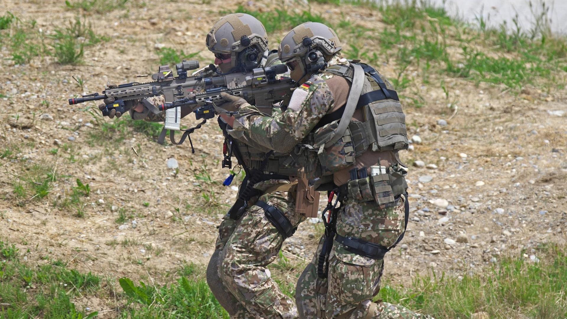 Zwei Soldaten des Kommandos Spezialkräfte in voller Kampfmontur und mit Machinengewehr im Anschlag pirschen sich einen Weg entlang.