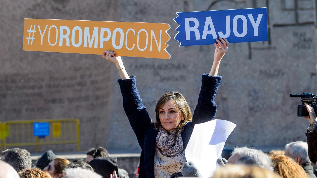 Frau sitzt auf den Schultern von jemandem in einer Menschenmenge und hält Schilder hoch, auf denen auf Spanisch steht: "Ich breche mit Rajoy."