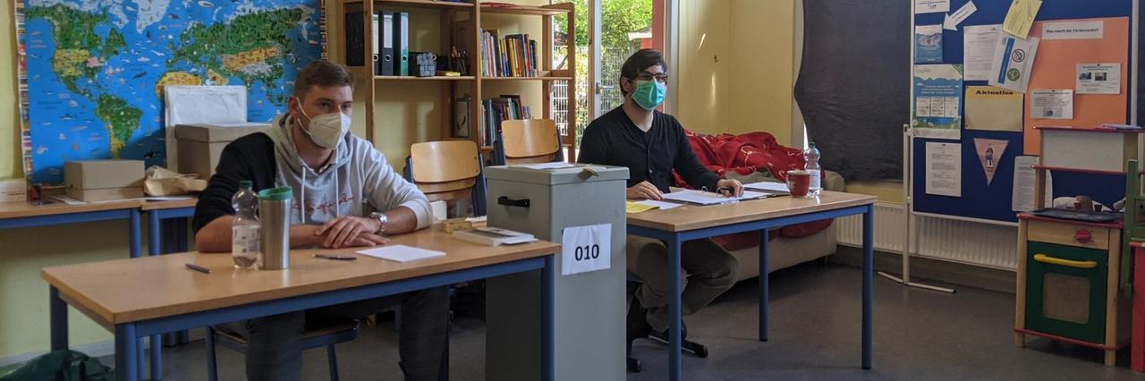 Zwei Wahl-Helfer sitzen an Schul-Tischen, zwischen ihnen steht die Wahl-Urne. Zwei Wahl-Helfer sitzen an Schul-Tischen, zwischen ihnen steht die Wahl-Urne.