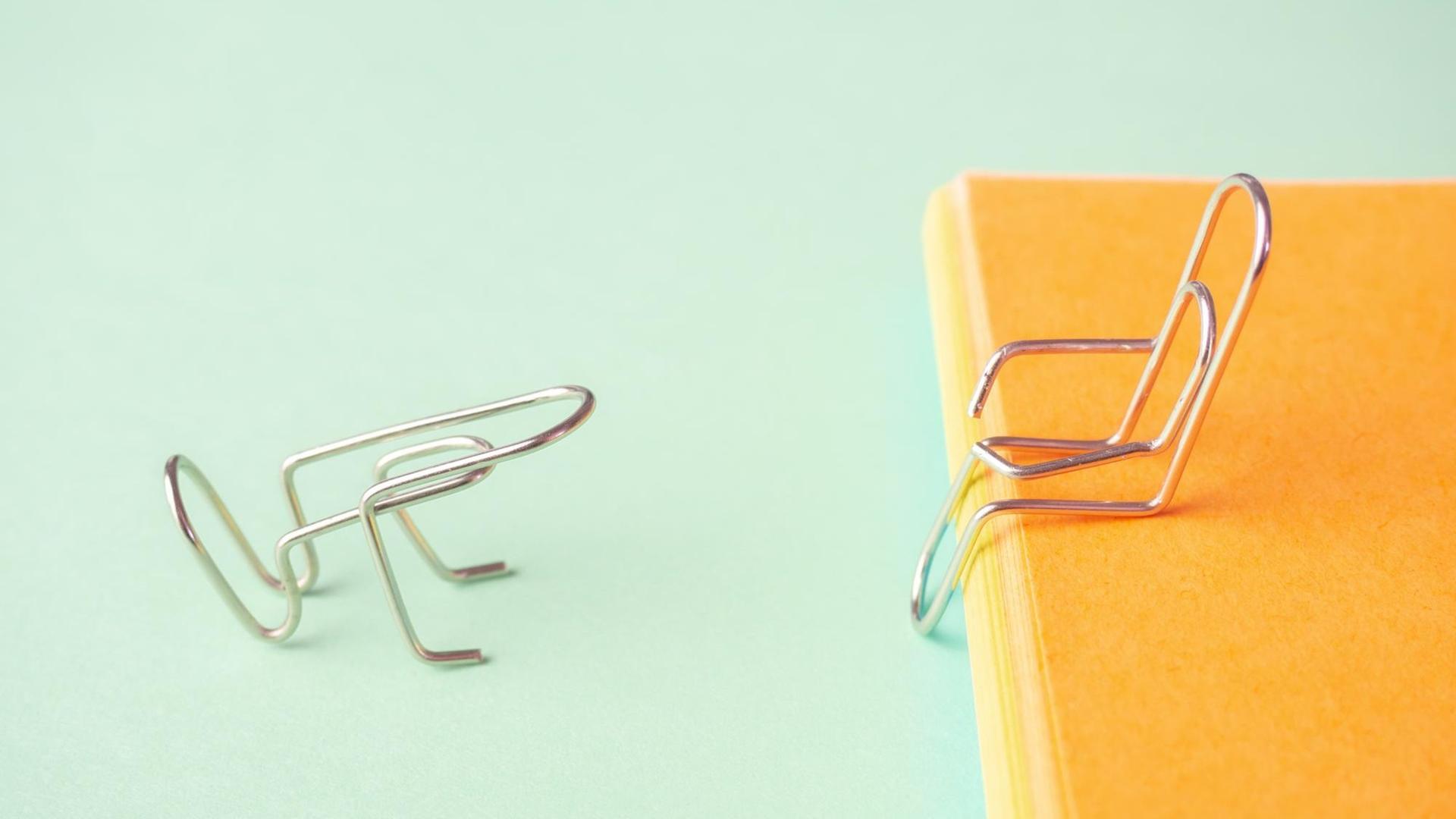 Auf pastellgrünem Hintergrund sind zwei Büroklammern zu sehen: sie sind so zurechtgebogen, dass die eine erhöht auf einer orangenen Fläche sitzt während die andere vor ihr kniet. Auf pastellgrünem Hintergrund sind zwei Büroklammern zu sehen: sie sind so zurechtgebogen, dass die eine erhöht auf einer orangenen Fläche sitzt während die andere vor ihr kniet.