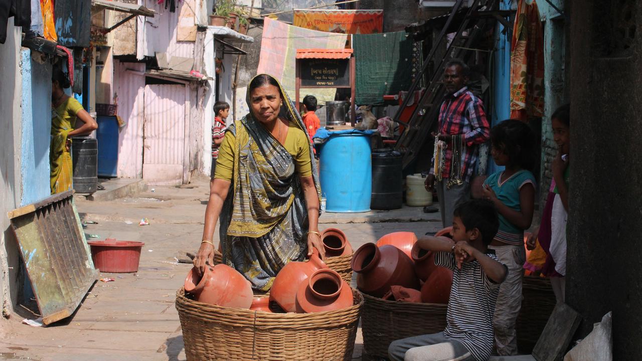 Ramiben Devaliya steht im Slum Dharavi hinter Krügen. Jeden Tag transportiert sie auf ihrem Kopf Körbe voller Krüge von Dharavi in einen Vorort Mumbais. Mitten in der Finanzmetropole Mumbai liegt Dharavi, einer der größten Slums Asiens.