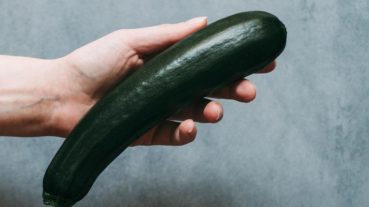 Eine Hand hält eine Salatgurke in der Hand. Die Wand im Hintergrund ist grau. Eine Hand hält eine Salatgurke in der Hand. Die Wand im Hintergrund ist grau.