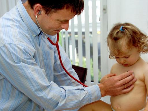 Ein kleines Mädchen sitzt mit skeptischer Mimik während der fälligen U7 Kinderuntersuchung (21. bis 24. Lebensmonat) beim Kinderarzt auf einem Behandlungstisch und wird mit dem Stethoskop abgehört.