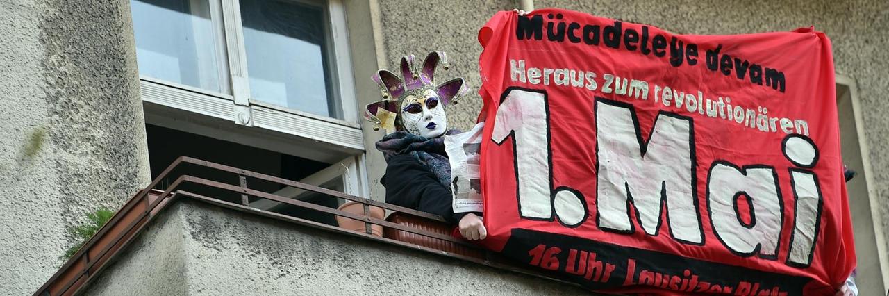 Ein maskierter Mensch hält ein Transparent mit der Aufschrift "Heraus zum revolutionären 1. Mai" von einem Balkon. 