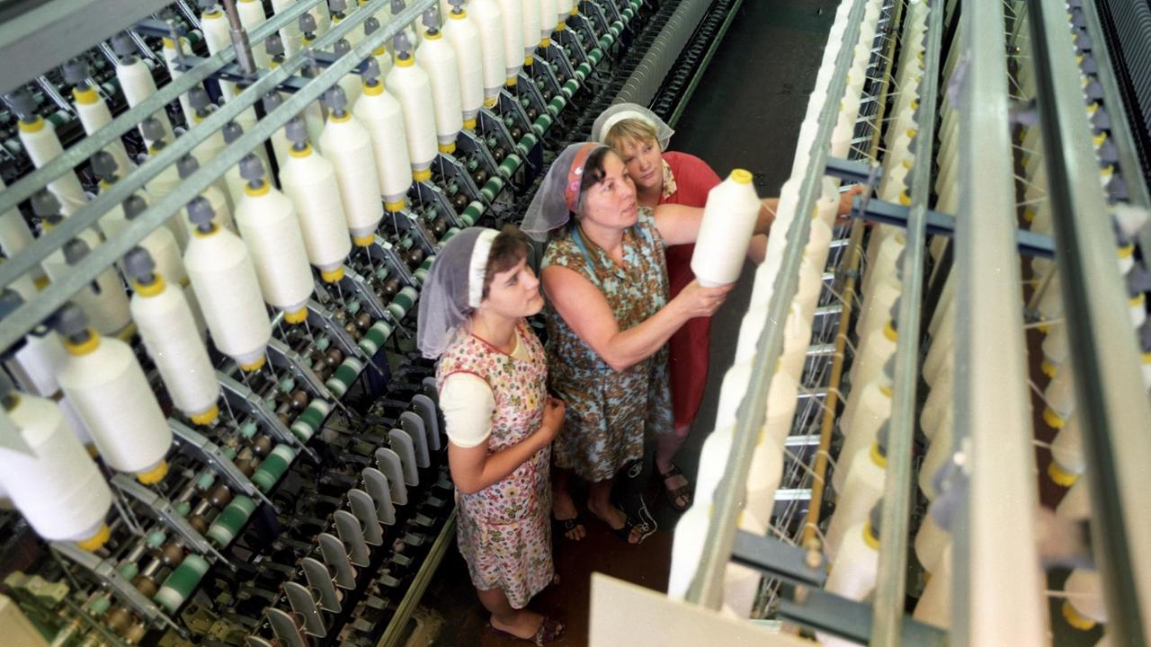 Drei Frauen mit Kittelschürzen und Kopftüchern stehen bei der Arbeit an einer Textilmaschine im VEB Kombinat Baumwolle in der DDR, aufgenommen in den 1980er Jahren Drei Frauen mit Kittelschürzen und Kopftüchern stehen bei der Arbeit an einer Textilmaschine im VEB Kombinat Baumwolle in der DDR, aufgenommen in den 1980er Jahren