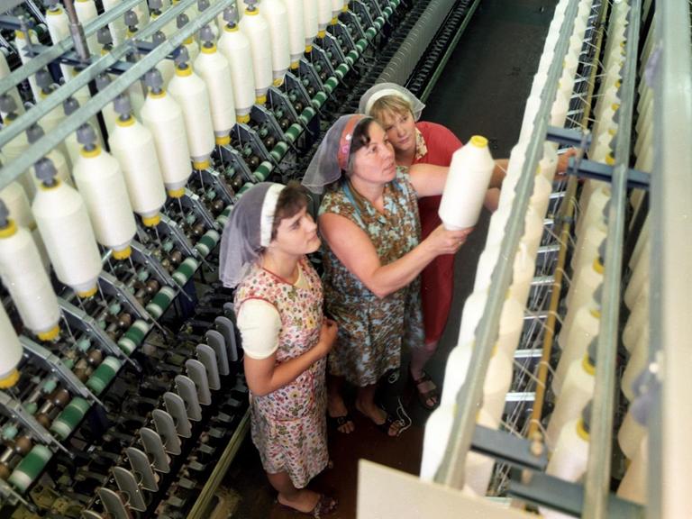 Drei Frauen mit Kittelschürzen und Kopftüchern stehen bei der Arbeit an einer Textilmaschine im VEB Kombinat Baumwolle in der DDR, aufgenommen in den 1980er Jahren