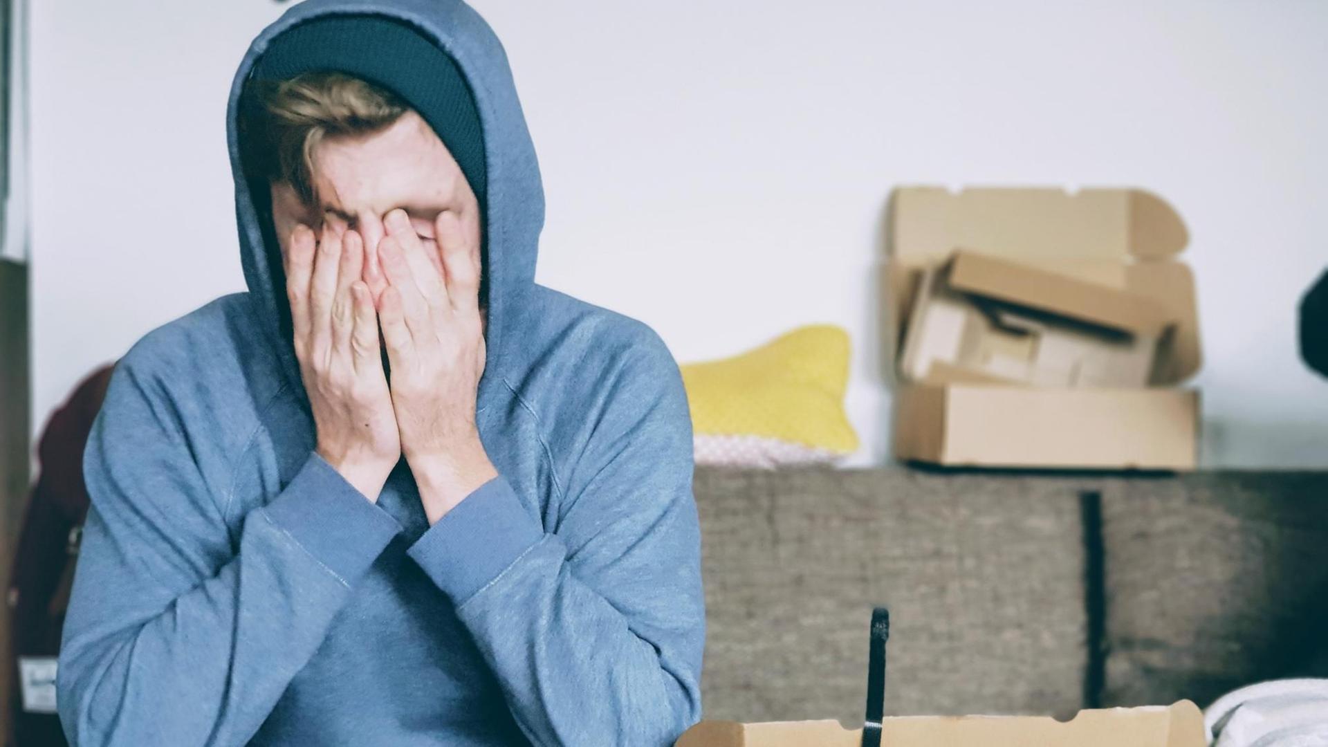 Eine Person mit Kapuzenpulli sitzt auf dem Sofa und hält sich beide Hände vor das Gesicht. Eine Person mit Kapuzenpulli sitzt auf dem Sofa und hält sich beide Hände vor das Gesicht.