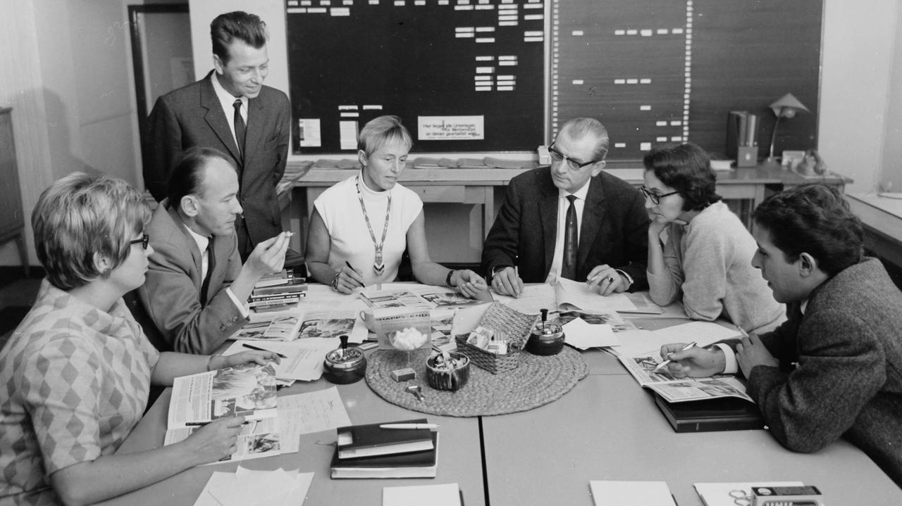 Ein historisches schwarz/weiss Foto von Beate Uhse in einer Besprechungssituation an einem Tisch mit Geschäftsleuten und Mitarbeitern. 