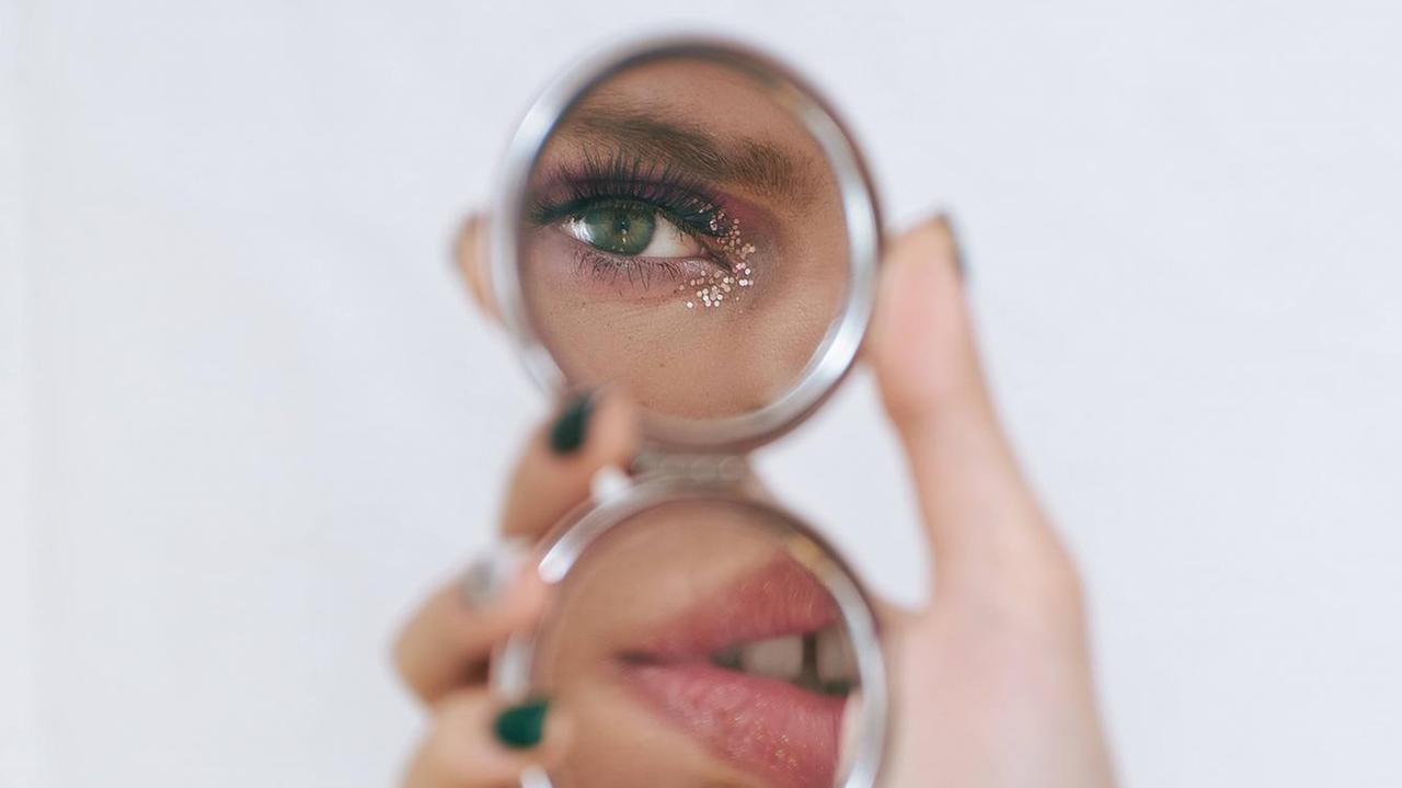 Ein Transmensch schaut sich in einem kleinen Handspiegel an, nur die geschminkten Augen und Lippen sind zu sehen. Ein Transmensch schaut sich in einem kleinen Handspiegel an, nur die geschminkten Augen und Lippen sind zu sehen.