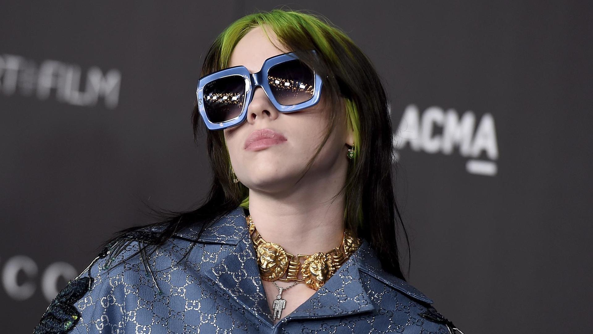 Billie Eilish posiert mit großer Sonnenbrille und grün-schwarzem Haar