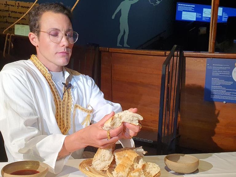 Der Schauspieler Brix Schaumburg in eimer Theaterszene. Er sitzt als queerer Jesus an einem Tisch und bricht Brot.