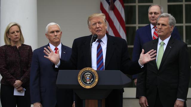 Trump steht an seinem Rednerpult, spricht und gestikuliert mit beiden Händen. Die anderen stehen schweigend hinter ihm und schauen mit ernstem Blick zu.