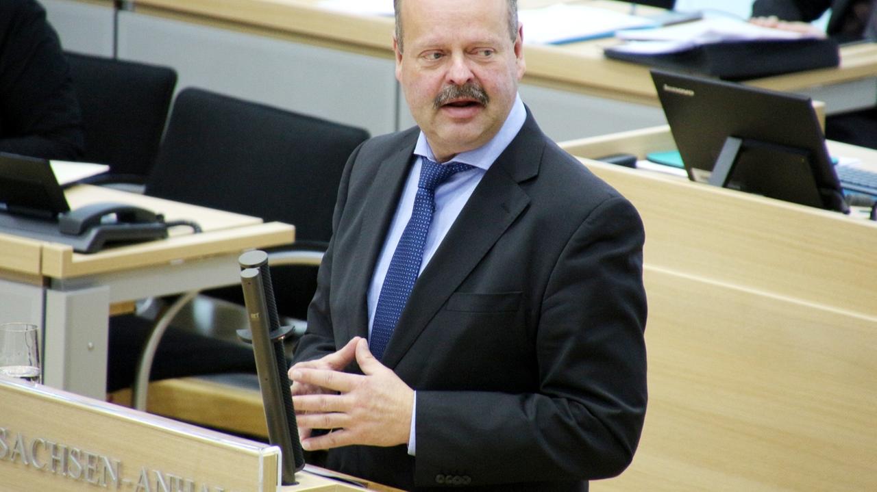 Ein Mann mit Schnurrbart steht an einem Rednerpult und blickt zur Seite.
