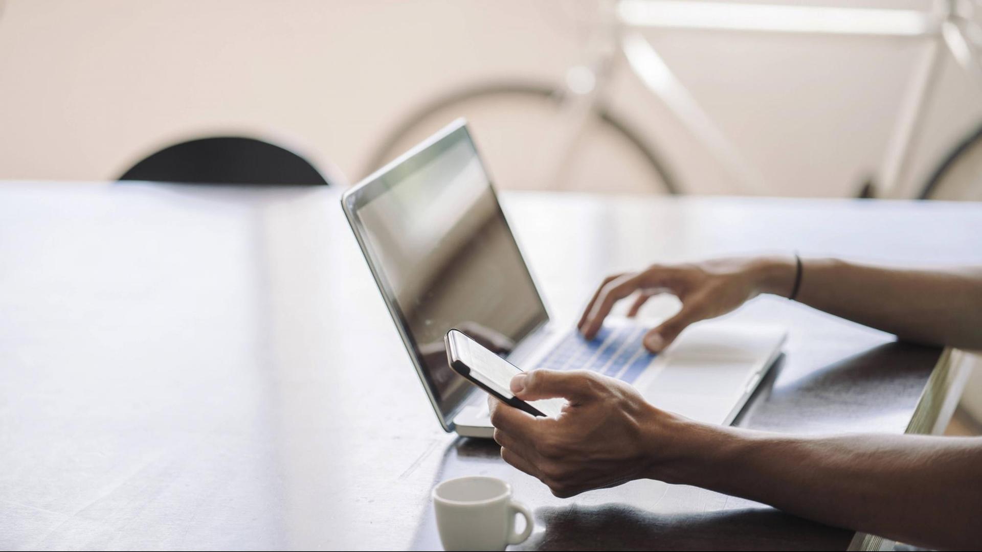 Jemand sitzt an einem Tisch vor einem Laptop und hält mit einer Hand ein Smartphone, im Hintergrund ist ein Fahrrad zu sehen, im Vordergrund eine Tasse. Jemand sitzt an einem Tisch vor einem Laptop und hält mit einer Hand ein Smartphone, im Hintergrund ist ein Fahrrad zu sehen, im Vordergrund eine Tasse.