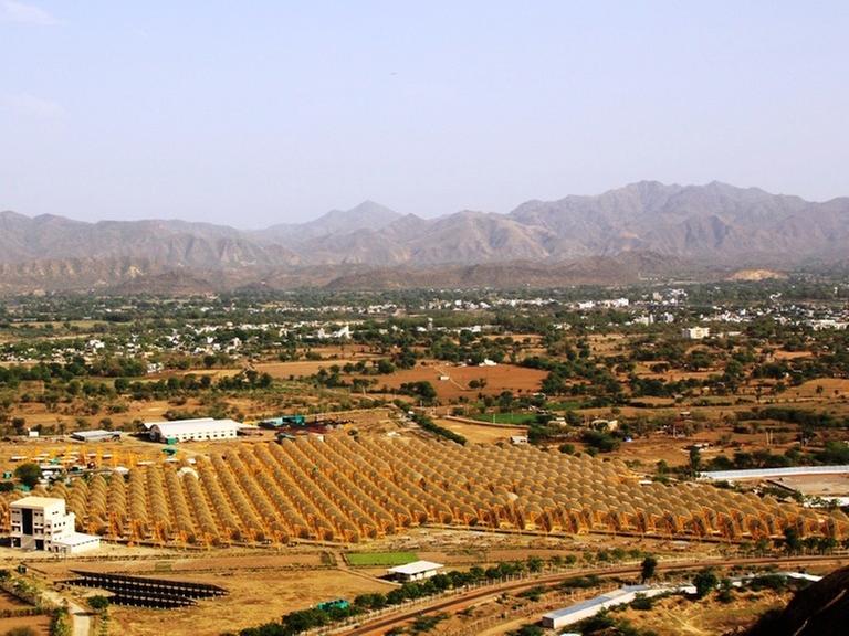 In einer Ebene von Rajasthan stehen zahlreiche Parabolschüsseln des solarthermischen Kraftwerks "India One". Im Hintergrund sind kahle Berge zu sehen.