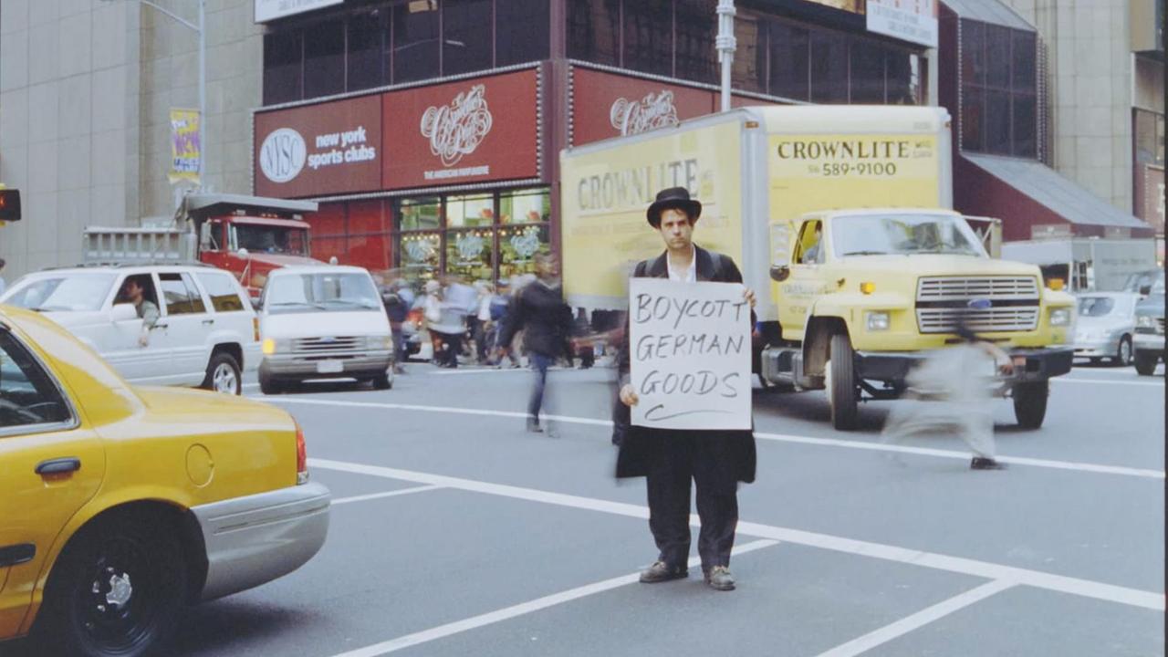 Szene aus dem Dokumentarfilm "Schlingensief - in das Schweigen hineinschreien": Christoph Schlingensief steht, als traditioneller Jude verkleidet, auf einer Straße in New York. Er hält ein Schild mit dem Aufdruck "Boycott German Goods". Szene aus dem Dokumentarfilm "Schlingensief - in das Schweigen hineinschreien": Christoph Schlingensief steht, als traditioneller Jude verkleidet, auf einer Straße in New York. Er hält ein Schild mit dem Aufdruck "Boycott German Goods".