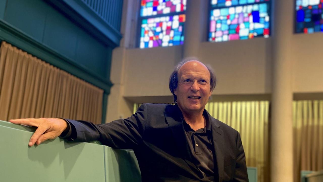 Der Dirigent sitzt in den leeren Kirchenbänken und lächelt in die Kamera. Der Dirigent sitzt in den leeren Kirchenbänken und lächelt in die Kamera.