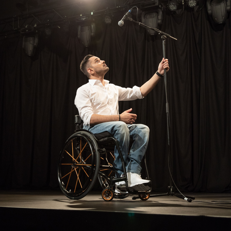 Tan Caglar sitzt in seinem Rollstuhl auf der Bühne und blickt hoch auf ein Mikrofon.
