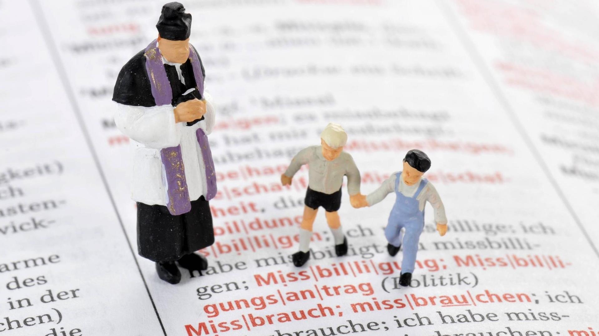 Modelleisenbahnfiguren eines Priesters und zweier Kinder auf einer aufgeschlagenen Wörterbuchseite vor dem Wort "Missbrauch" Modelleisenbahnfiguren eines Priesters und zweier Kinder auf einer aufgeschlagenen Wörterbuchseite vor dem Wort "Missbrauch"