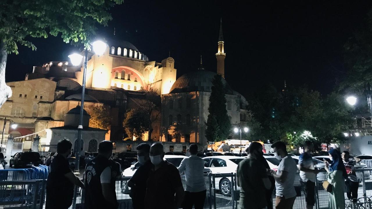 Mehrere Männer stehen im Dunklen vor der hell erleuchteten Hagia Sophia vor Absperrungen.