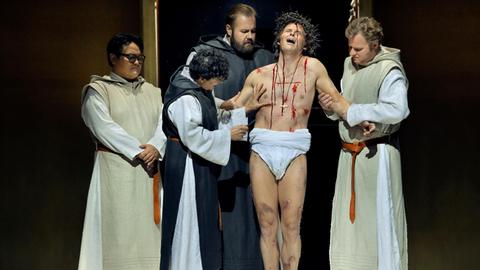 Eine Gruppe von Männern auf einer Theaterbühne stehen um einen aus einzelnen Wunden blutenden Mann herum. Die Operlamm-Szene.