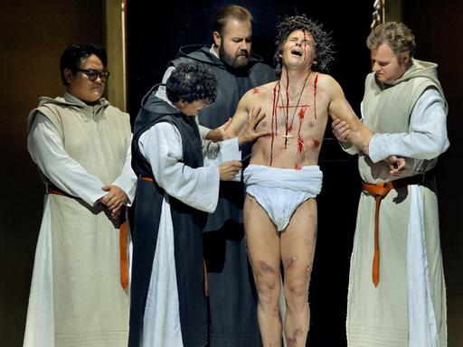 Eine Gruppe von Männern auf einer Theaterbühne stehen um einen aus einzelnen Wunden blutenden Mann herum. Die Operlamm-Szene.