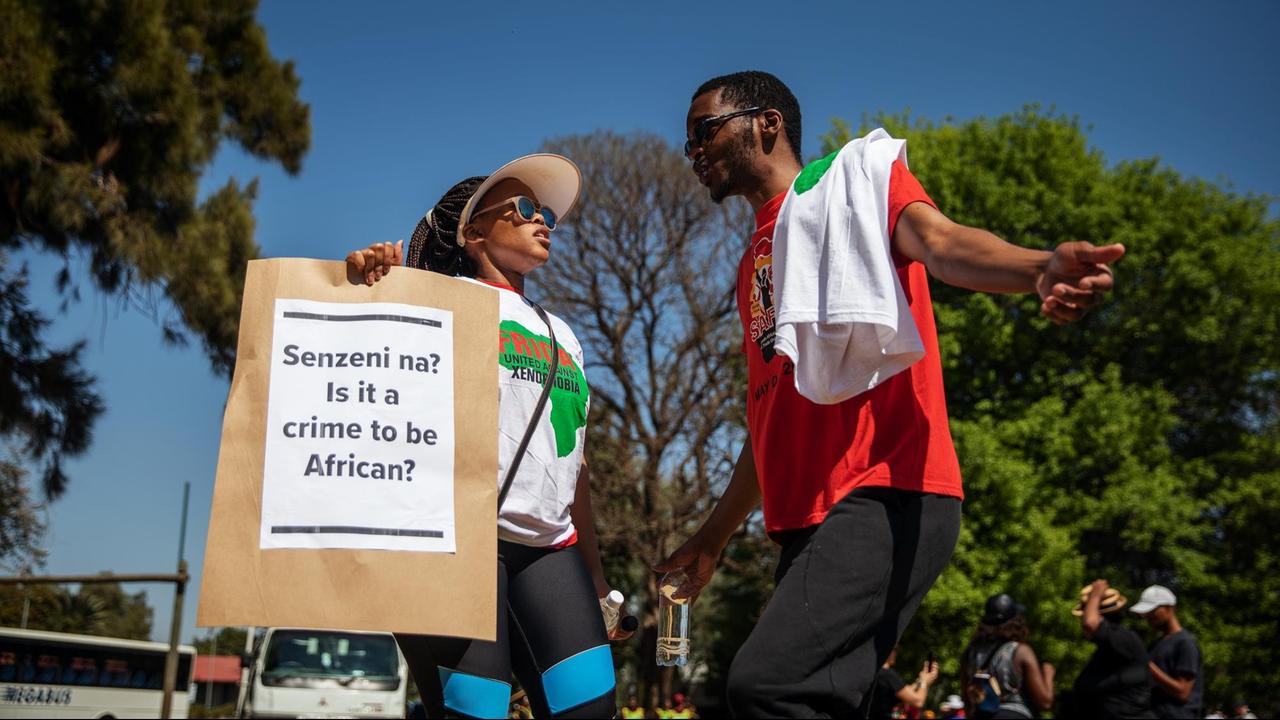Zwei Demonstranten tanzen und halten ein Schild mit der Aufstand: "Is it a crime to be African?" Zwei Demonstranten tanzen und halten ein Schild mit der Aufstand: "Is it a crime to be African?"