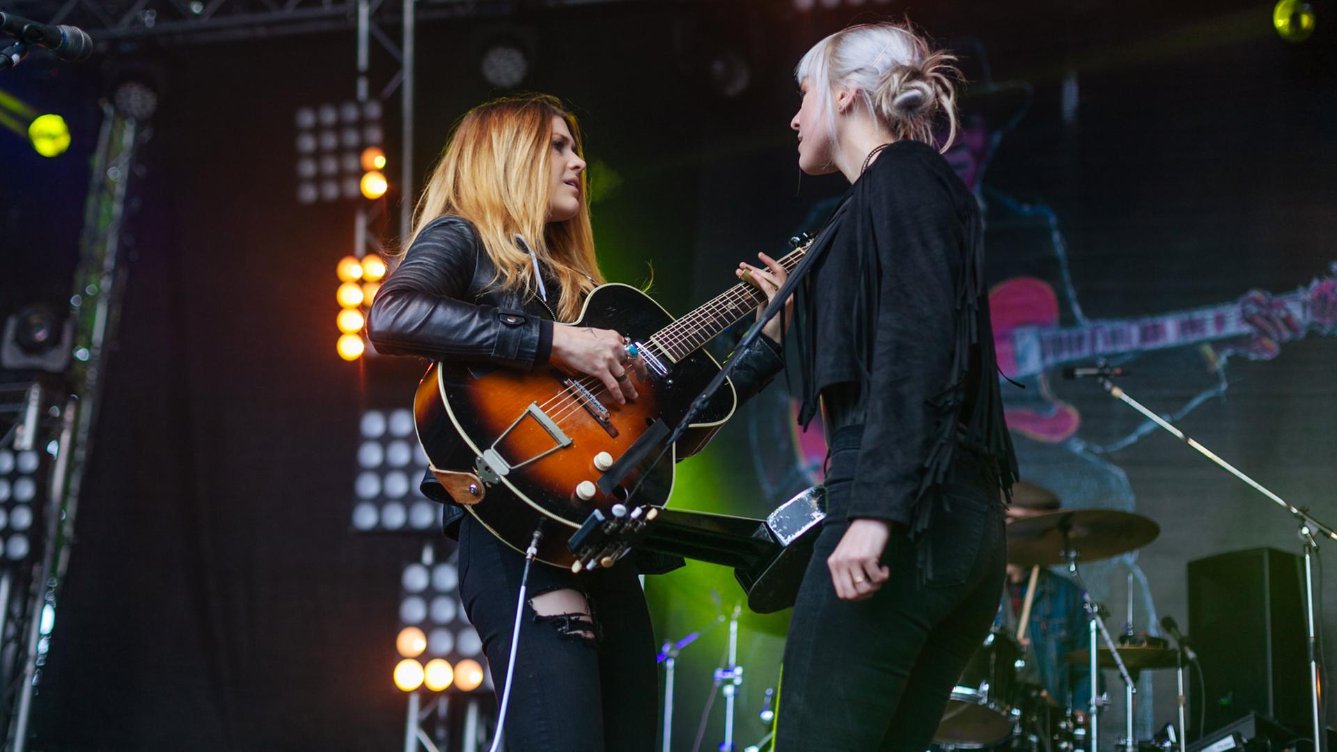 Zwei Frauen mit Gitarren stehen sich auf einer Konzertbühne gegenüber. Zwei Frauen mit Gitarren stehen sich auf einer Konzertbühne gegenüber.