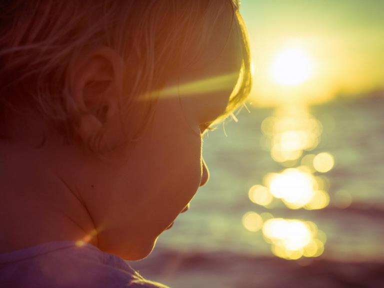 Fotografie eines Kindes am Meer, im Gegenlicht der, am Horizont, untergehenden Sonne.