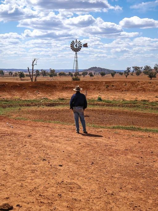 Ein australischer Bauer blickt auf ein Windrad, das einsam in der ausgedörrten Landschaft steht.